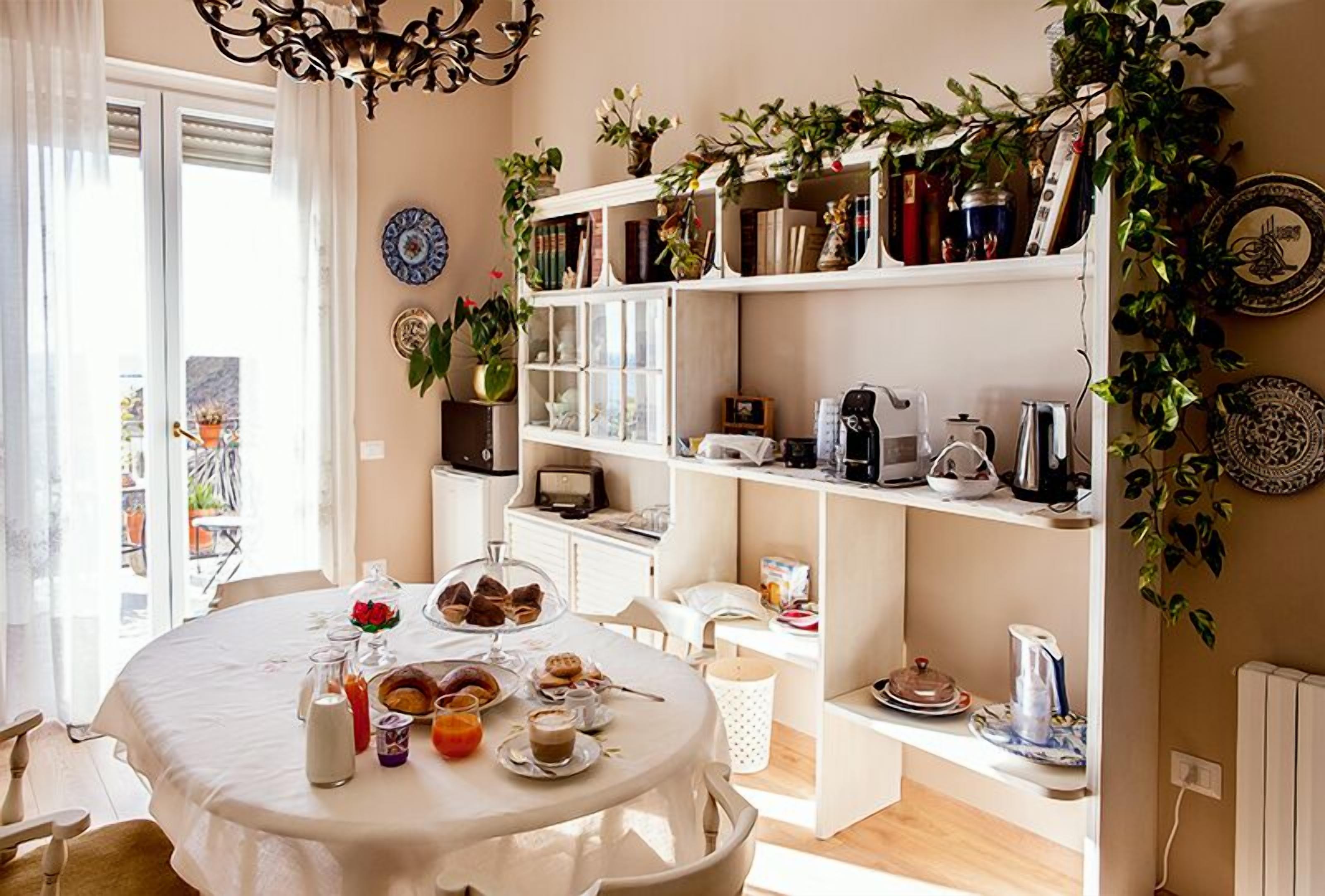 Une table de petit-déjeuner avec des viennoiseries et des boissons, une bibliothèque remplie de livres et de plantes, et une vue sur l’extérieur.