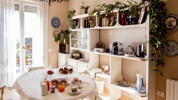 Une table de petit-déjeuner avec des viennoiseries et des boissons, une bibliothèque remplie de livres et de plantes, et une vue sur l’extérieur.