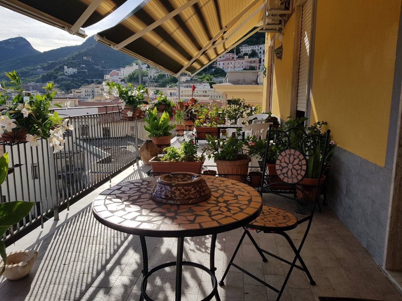 A balcony with a round table and two chairs, surrounded by potted plants and a decorative bowl.