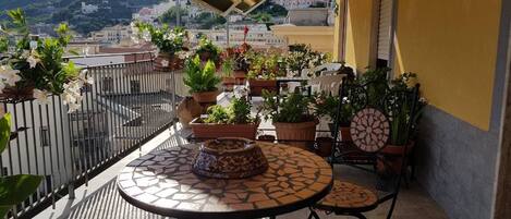 A balcony with a round table and two chairs, surrounded by potted plants and a decorative bowl.