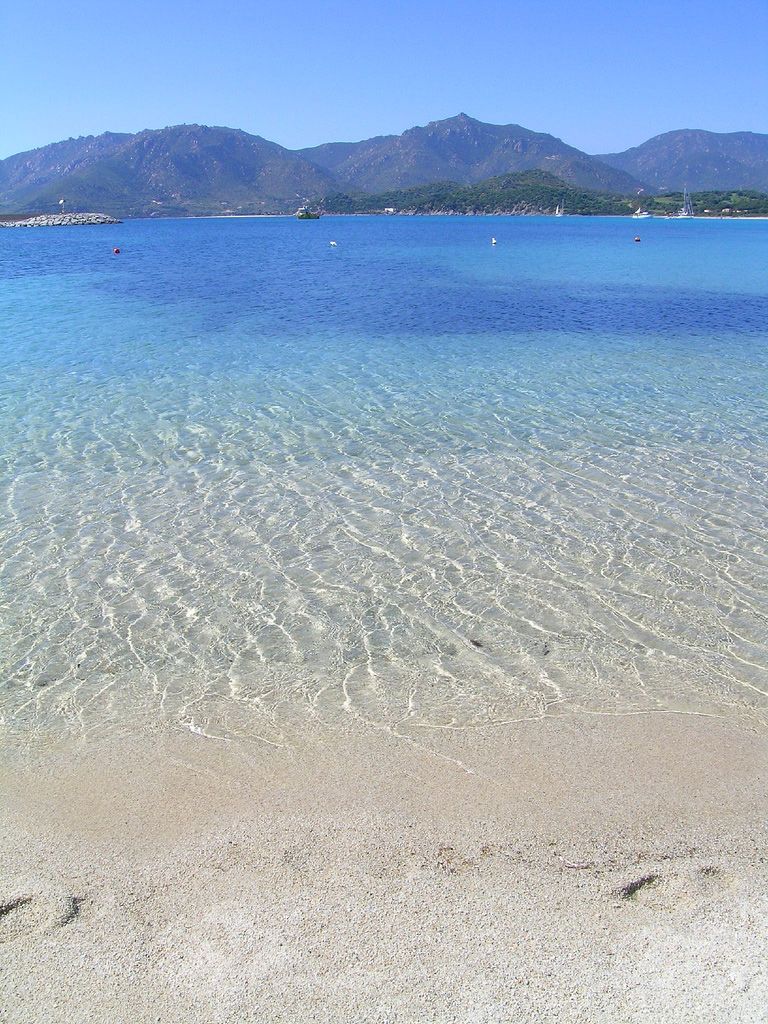 Une plage dégagée, avec du sable visible et une eau peu profonde, des montagnes en arrière-plan et quelques bateaux.