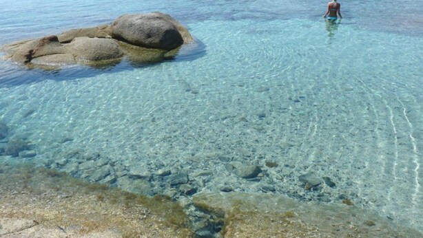 Une zone côtière dégagée, avec une grande formation rocheuse au premier plan et une personne debout dans l’eau.