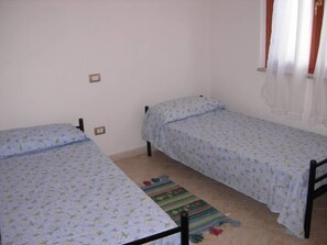 A small, simple room with two single beds, a colorful rug, and a plain wall with a window.