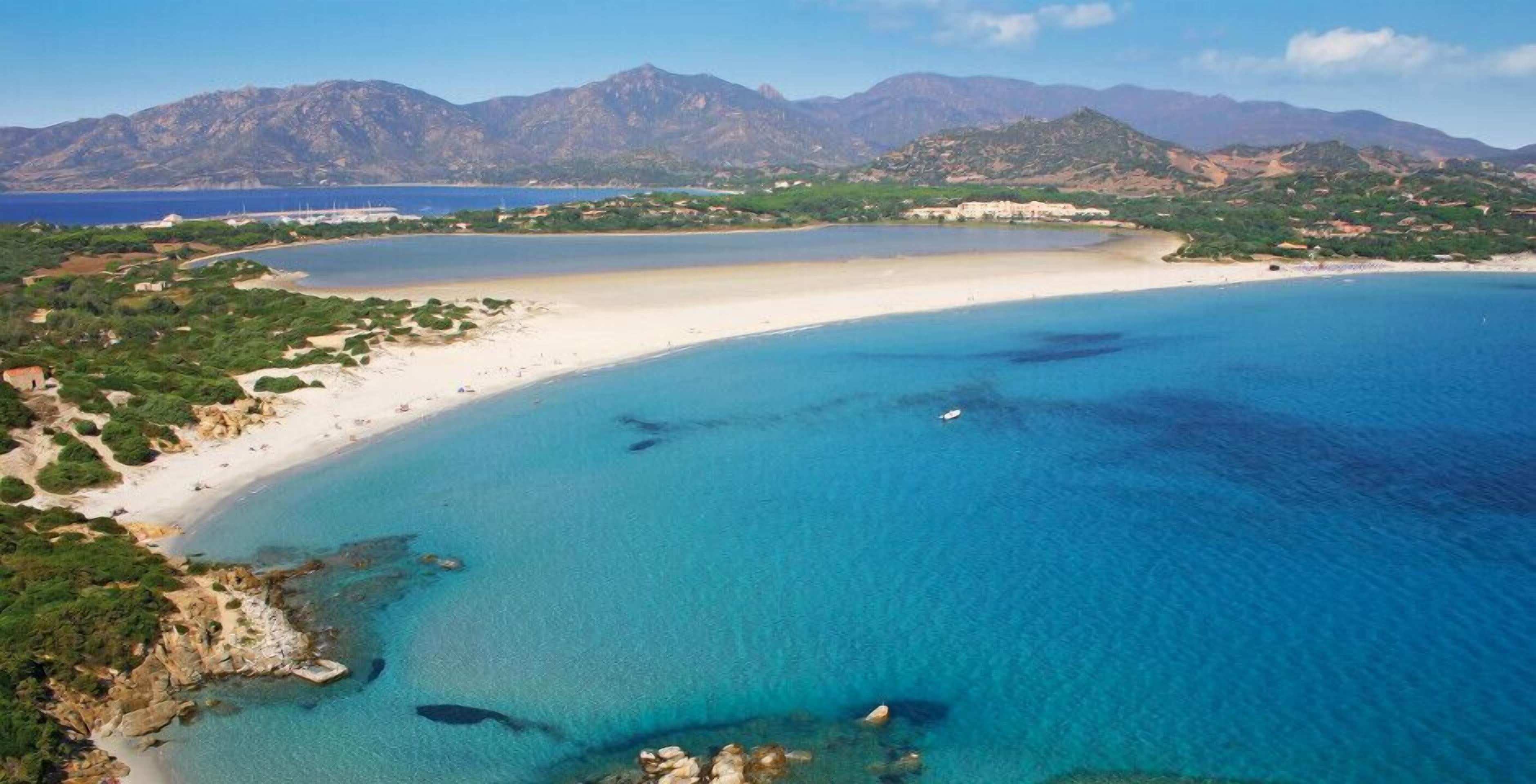 Vue aérienne d’une zone côtière avec une plage de sable, des eaux d’un bleu limpide et un paysage montagneux en arrière-plan.