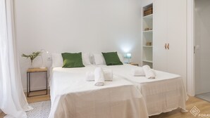 Ein Hotelzimmer mit zwei Betten, weißer Bettwäsche, grünen Kissen, einem Nachttisch mit Lampe, einer kleinen Pflanze und einem Schrank mit Regalen.