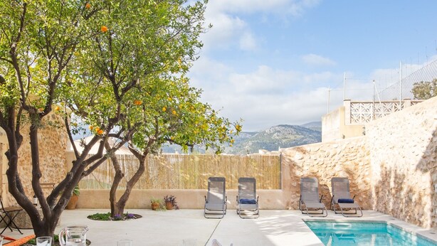 Ein Essbereich am Pool mit einem Tisch für zwei Personen, umgeben von Liegestühlen, einem Baum mit orangen Blüten und Blick auf Berge.