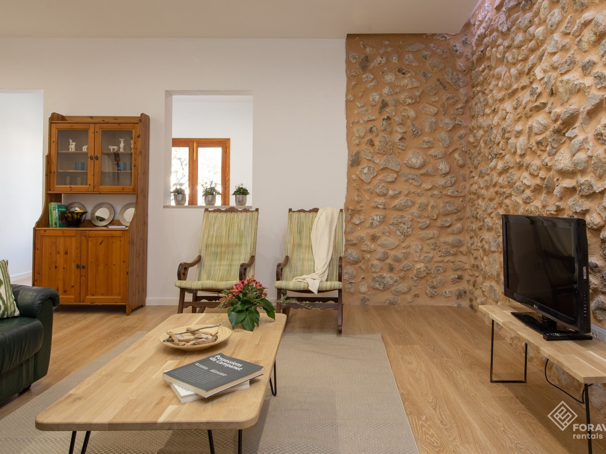A cozy living room with a wooden coffee table, two armchairs, a sofa, a stone wall, a wooden cabinet, and a television on a stand.