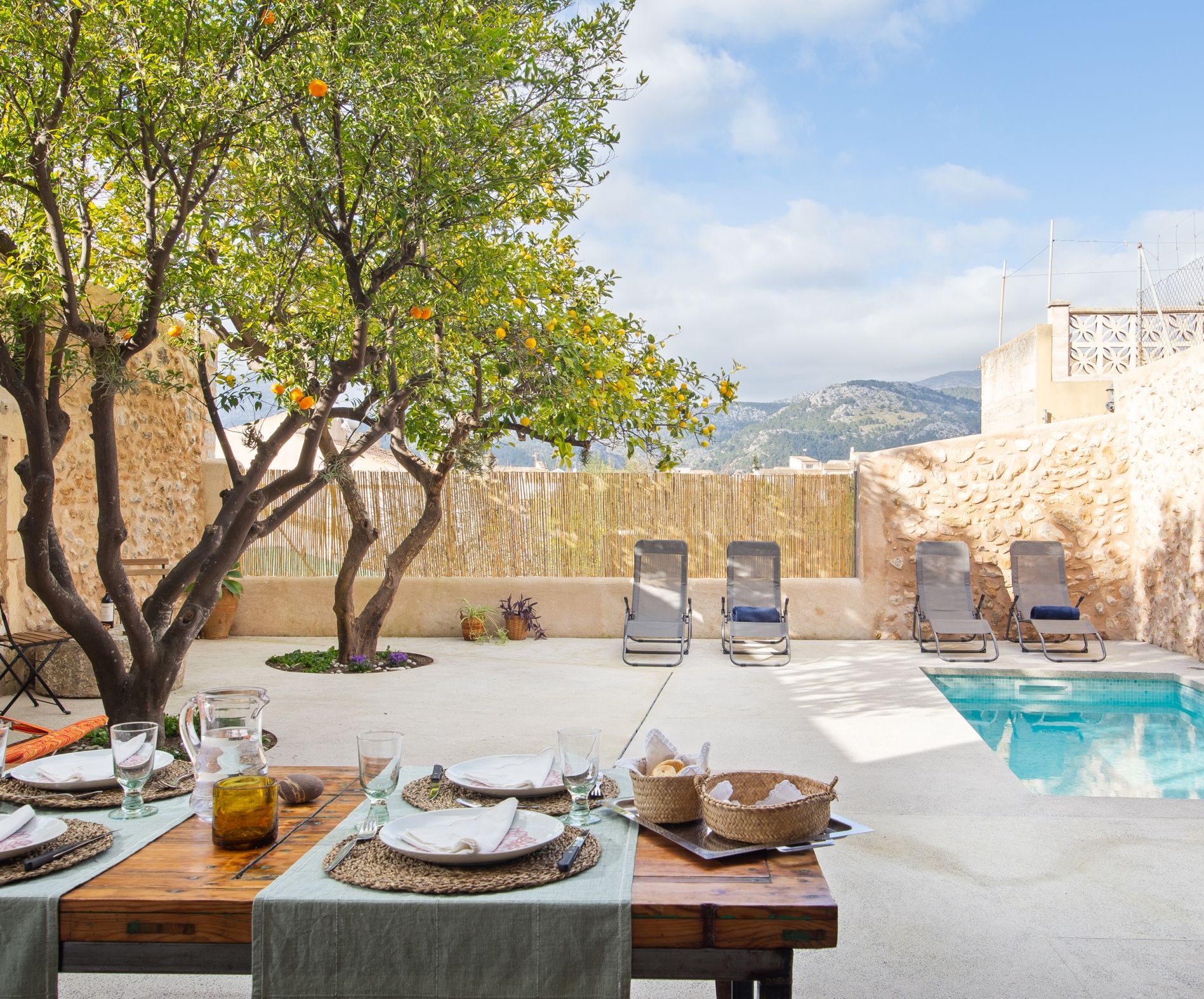 A poolside dining area with a table set for two, surrounded by lounge chairs, a tree with orange flowers, and a view of mountains.