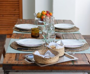 Une table en bois, mise pour un repas, avec des assiettes, des verres et un panier de fruits en arrière-plan.