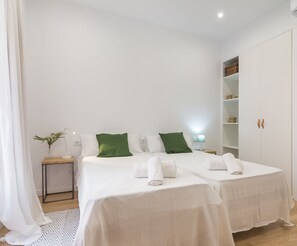 Une chambre d’hôtel avec deux lits, du linge de lit blanc, des oreillers verts, une table de chevet avec une lampe, une petite plante et une armoire avec des étagères.