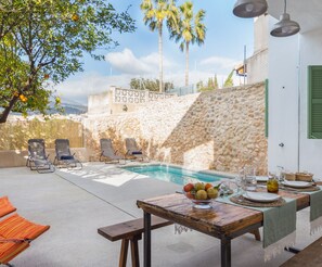 Un espace aménagé au bord de la piscine, avec des chaises longues, une table en bois sur laquelle sont disposés des fruits et un mur en pierre.