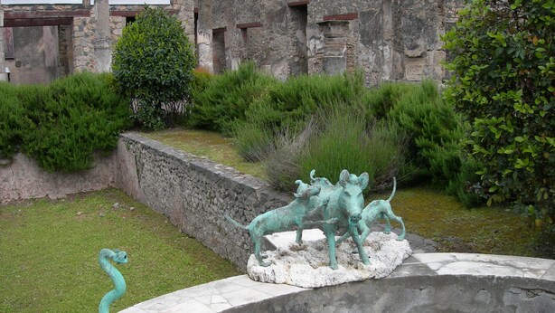 Ein Garten mit Steinmauer, einem kleinen Teich und einer Bronzefigur von drei Tieren.