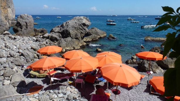 Una playa rocosa con sombrillas naranjas y gente relajándose.