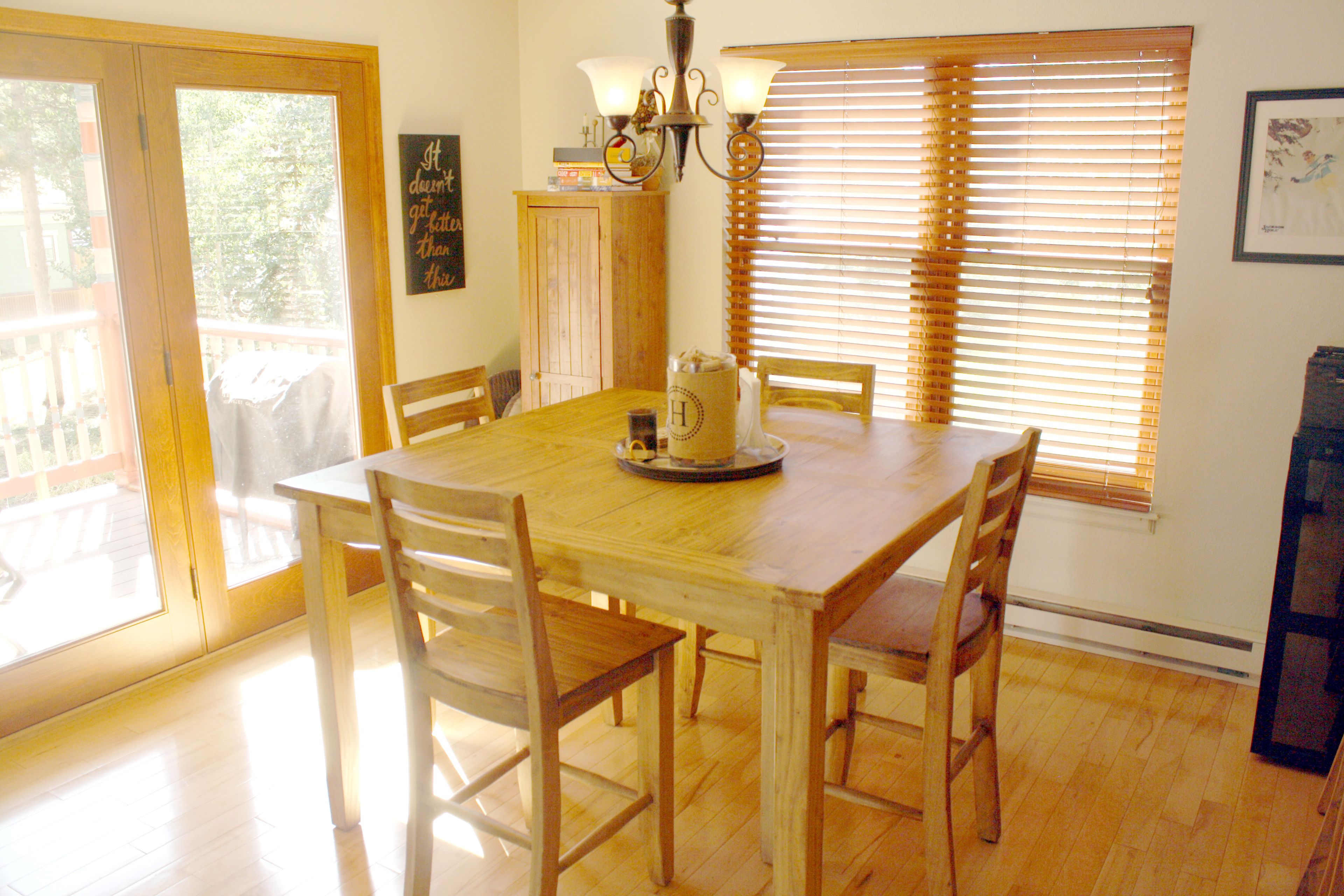 A dining area with a wooden table, four chairs, a chandelier, and a window with blinds.