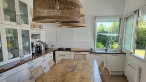 A modern kitchen with a wooden dining table, white cabinets, and a large window with a view of greenery. - ANDERNOS, 8 PERSONS VILLA 140M2 PETS WELCOME (Andernos-Les-Bains)