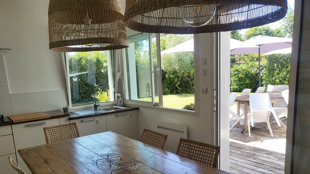 A kitchen with a wooden table, wicker chairs, and a view of an outdoor patio with white chairs and umbrellas. - ANDERNOS, 8 PERSONS VILLA 140M2 PETS WELCOME (Andernos-Les-Bains)