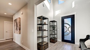 A spacious hallway with wooden flooring, a black-framed door, and two black metal shelving units with decorative items. - Zane's Place (St. George)