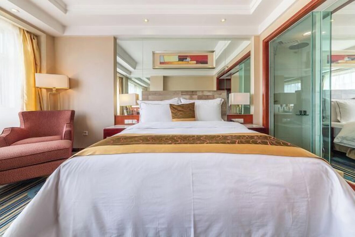 A hotel room with a large bed, a chair, a desk, and a glass partition.