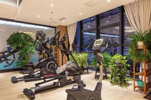 Une salle de sport équipée de vélos d’appartement et d’un tapis de course, de grandes fenêtres avec des rideaux et de plantes d’intérieur.