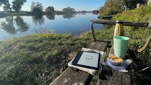 A serene lakeside picnic setup with a tablet displaying a smiley face, a thermos, a mug, and a container of food on a wooden bench. - Apartment in the nature and star park Westhavelland (Havelaue OT Wolsier)