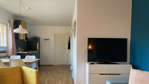 Un salon moderne comprenant un canapé, une table basse en verre, un téléviseur posé sur un meuble blanc, et un coin cuisine avec une table à manger et des chaises.