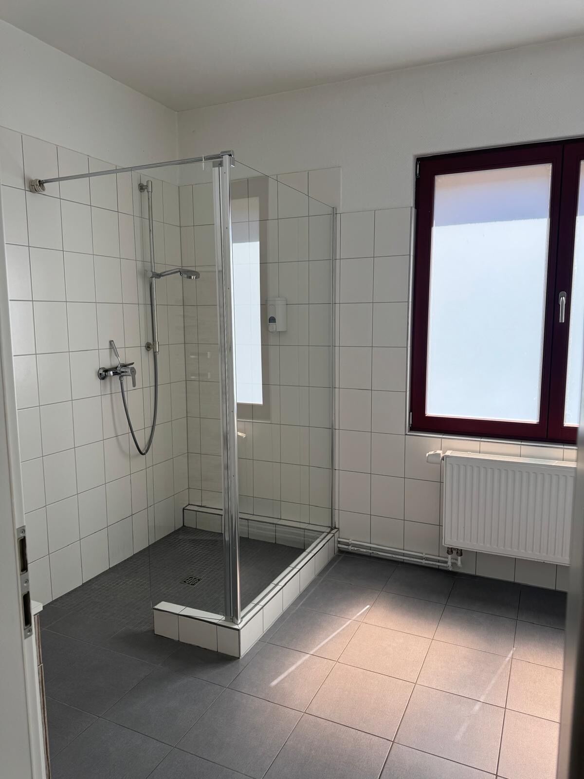 Eine Dusche mit Glastür, eine weiße Fliesenwand und ein Fenster mit dunklem Rahmen.