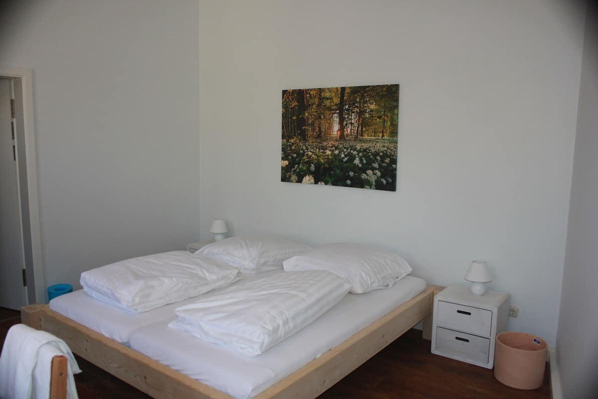 A single bed with white bedding, a nightstand with a lamp, and a picture of a forest on the wall.