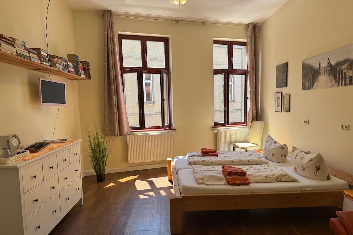 A bedroom with a bed, a dresser, a shelf with books, two windows, and a radiator.