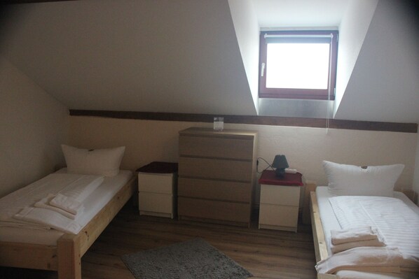 A small, dimly lit room with two single beds, a wooden dresser, and a small window.