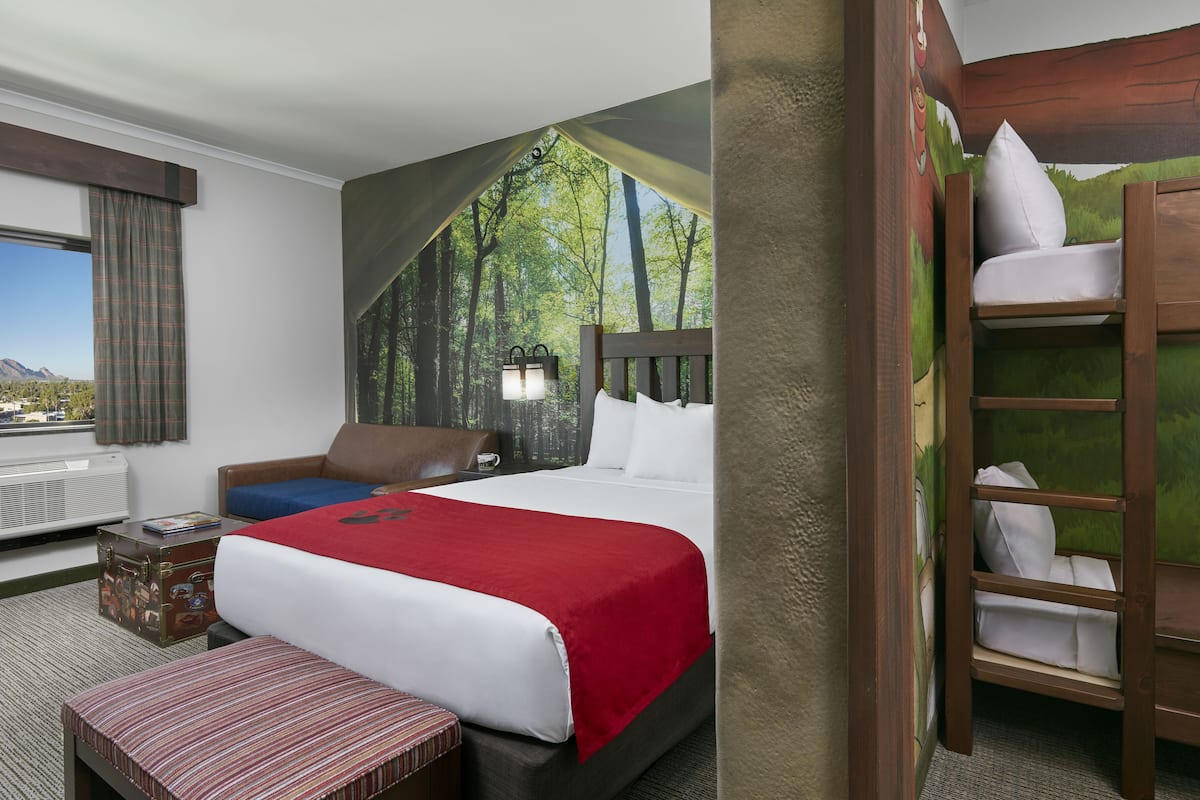 A hotel room with a bed, a bench, a suitcase, a window with a view, and a bunk bed.