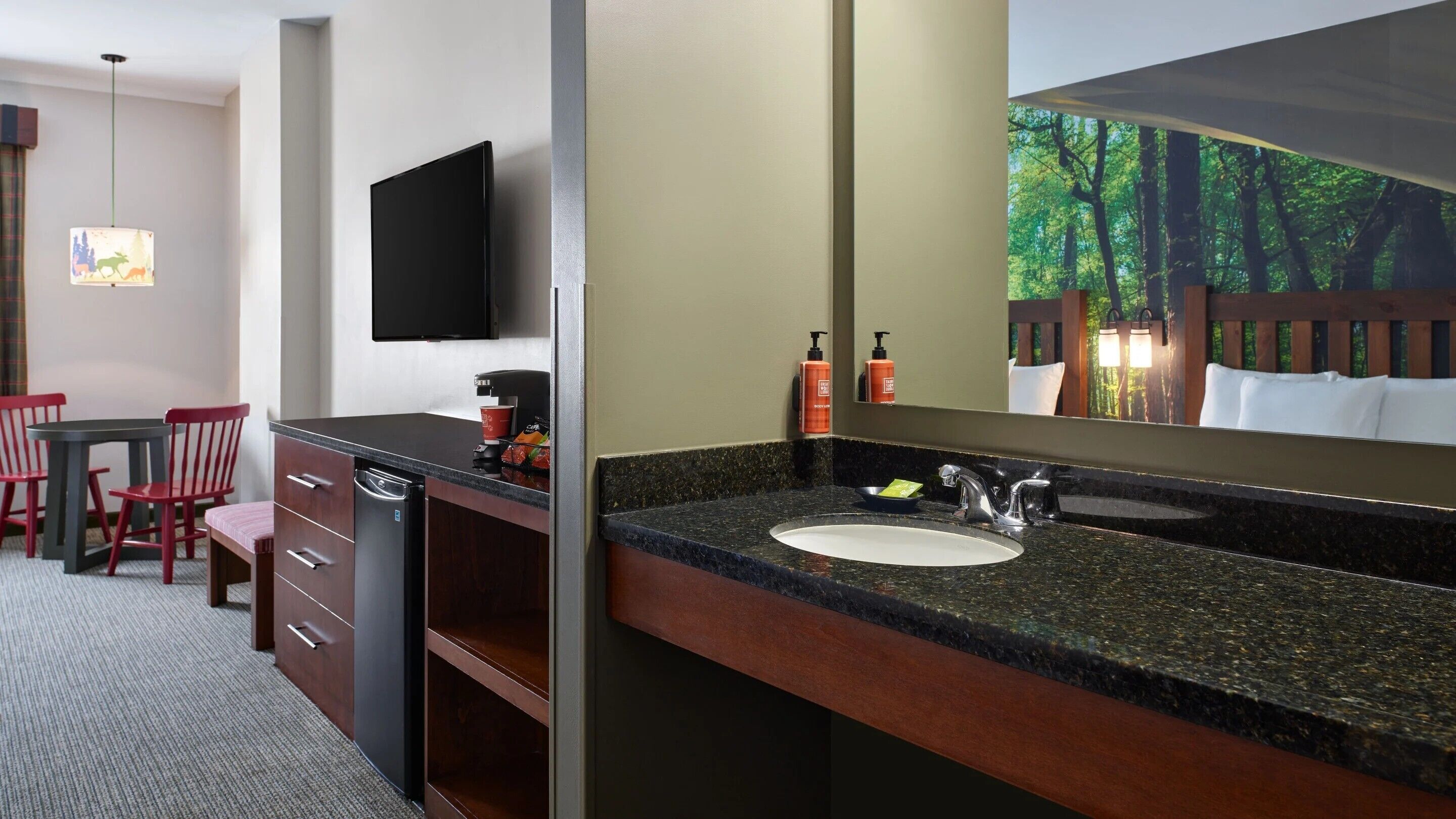 A hotel room with a kitchen area, a dining table with chairs, a sink with a mirror, and a flat-screen TV mounted on the wall.