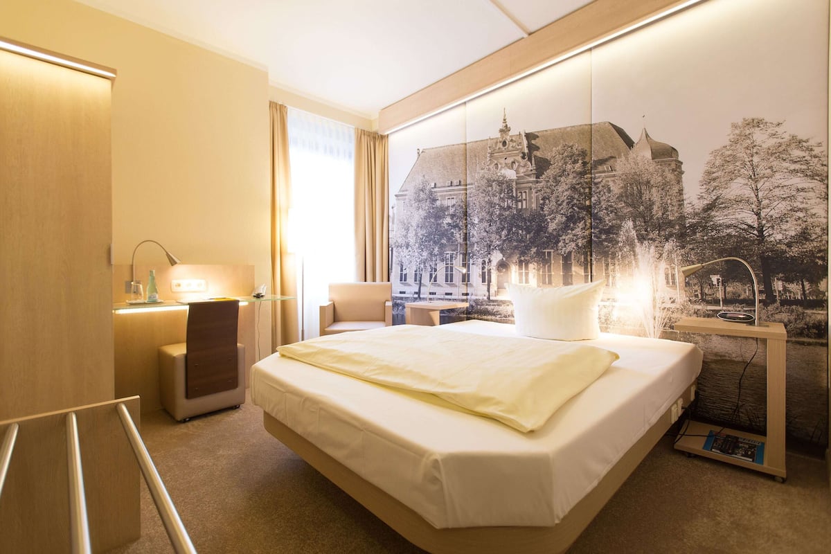A hotel room with a bed, a desk, a chair, a lamp, and a wall-mounted TV.