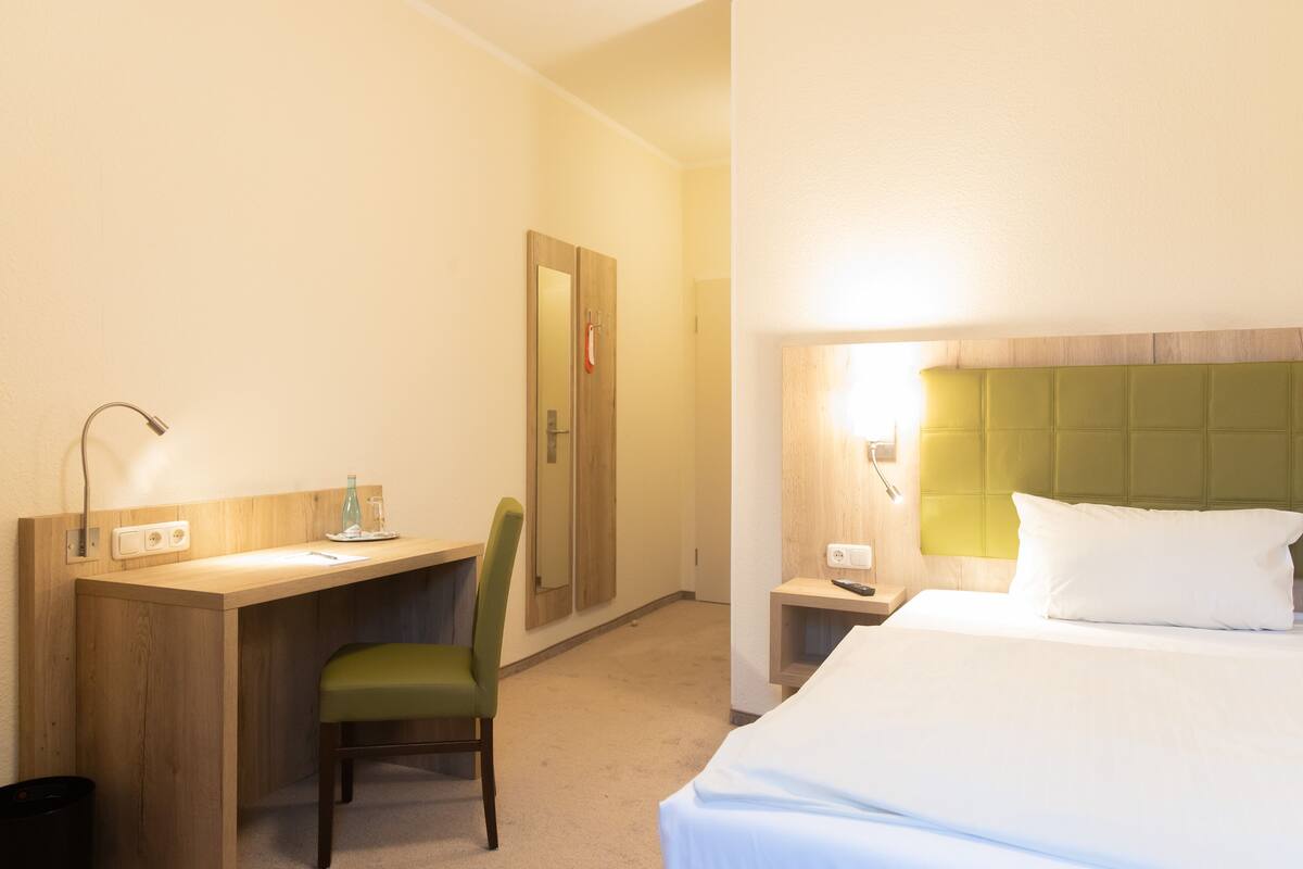 A hotel room with a bed, a desk, a chair, a nightstand, and a mirror.