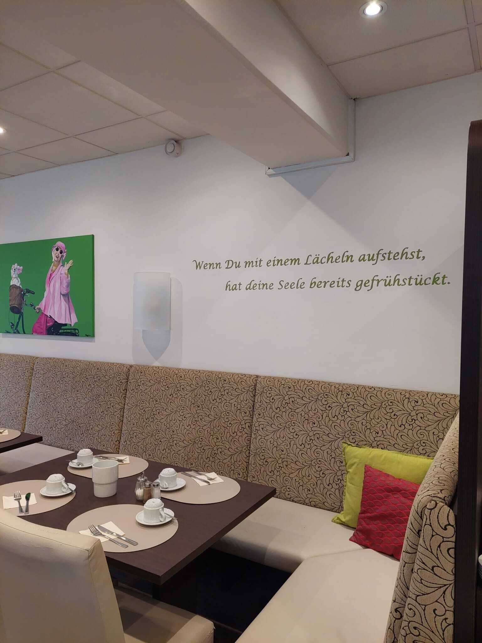 Uma sala de jantar com uma mesa posta para dois, uma parede com uma citação em alemão e um quadro verde acima da mesa.
