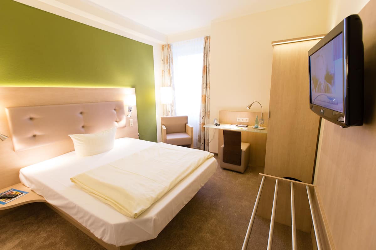 A hotel room with a large bed, a television, a desk with a lamp, and a chair.