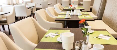 Un comedor de hotel con varias mesas dispuestas, vajilla blanca, manteles individuales verdes y un florero con una flor roja.
