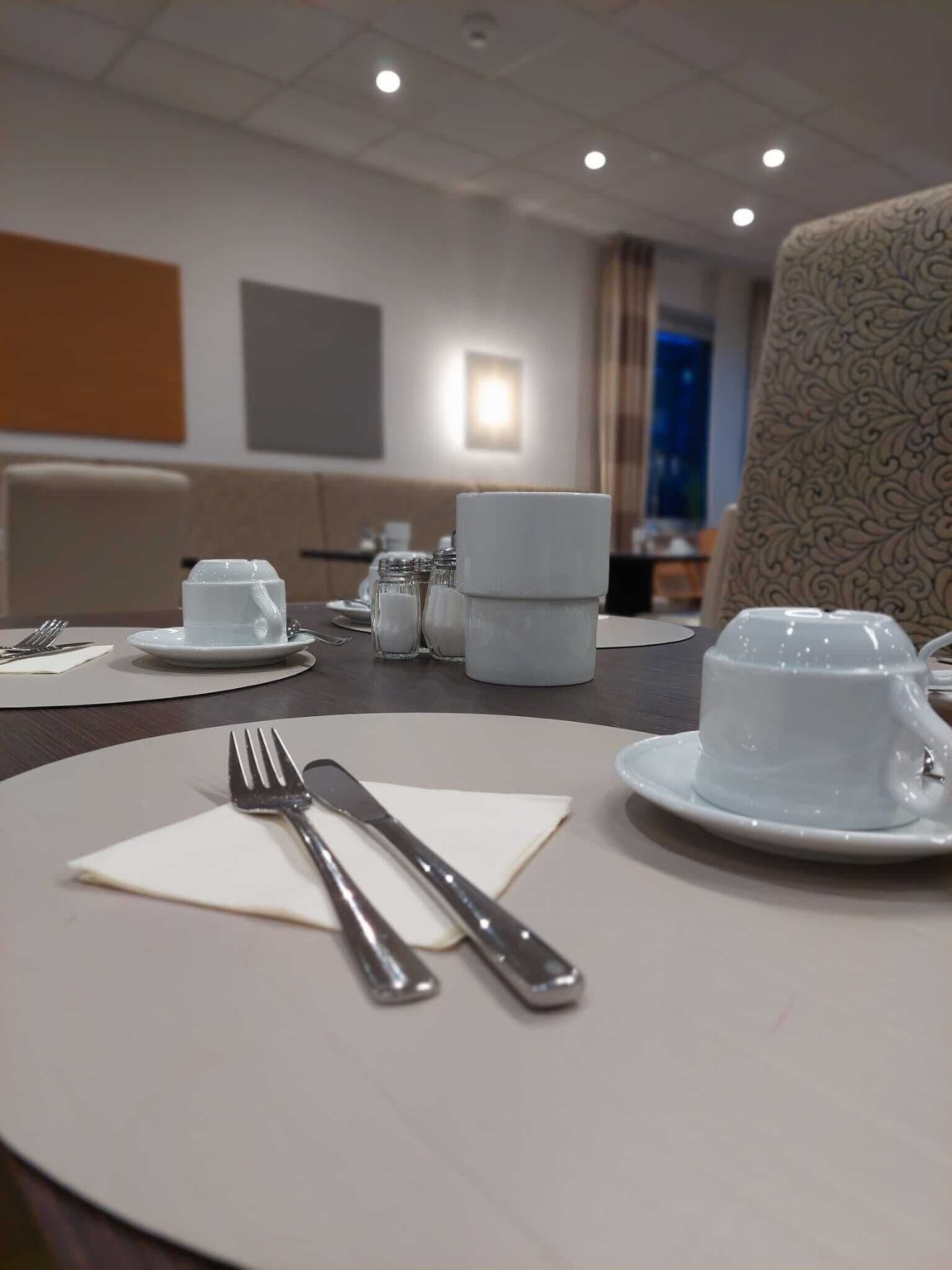 A hotel dining area with a table set for two, including plates, cutlery, and a cup.