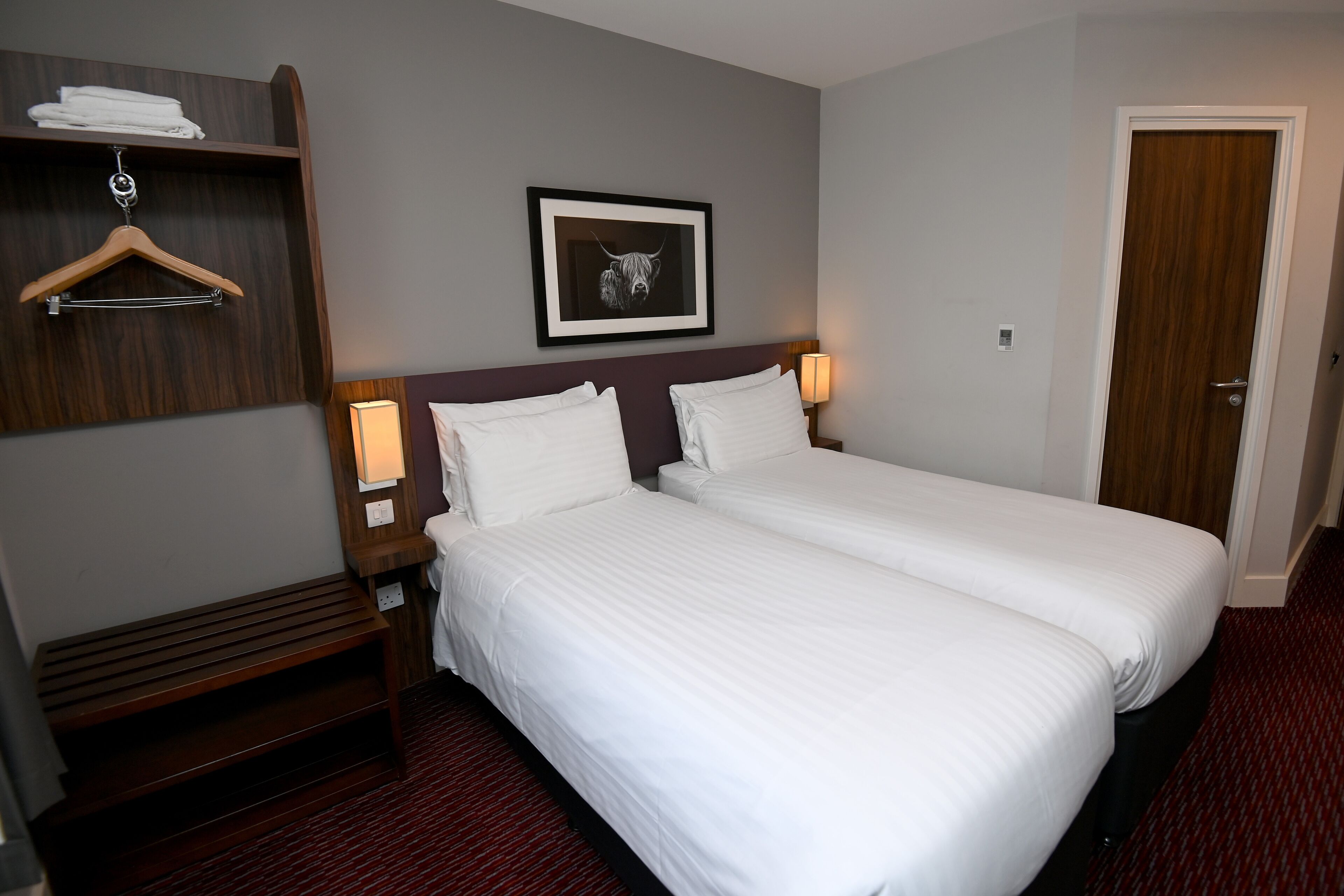 A hotel room with two single beds, a wooden bench, a coat rack, a framed picture on the wall, and a door.