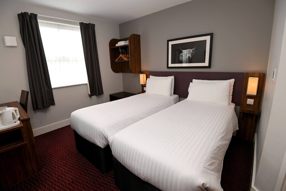 A hotel room with two beds, a wardrobe, a desk with a kettle, and a framed picture on the wall.