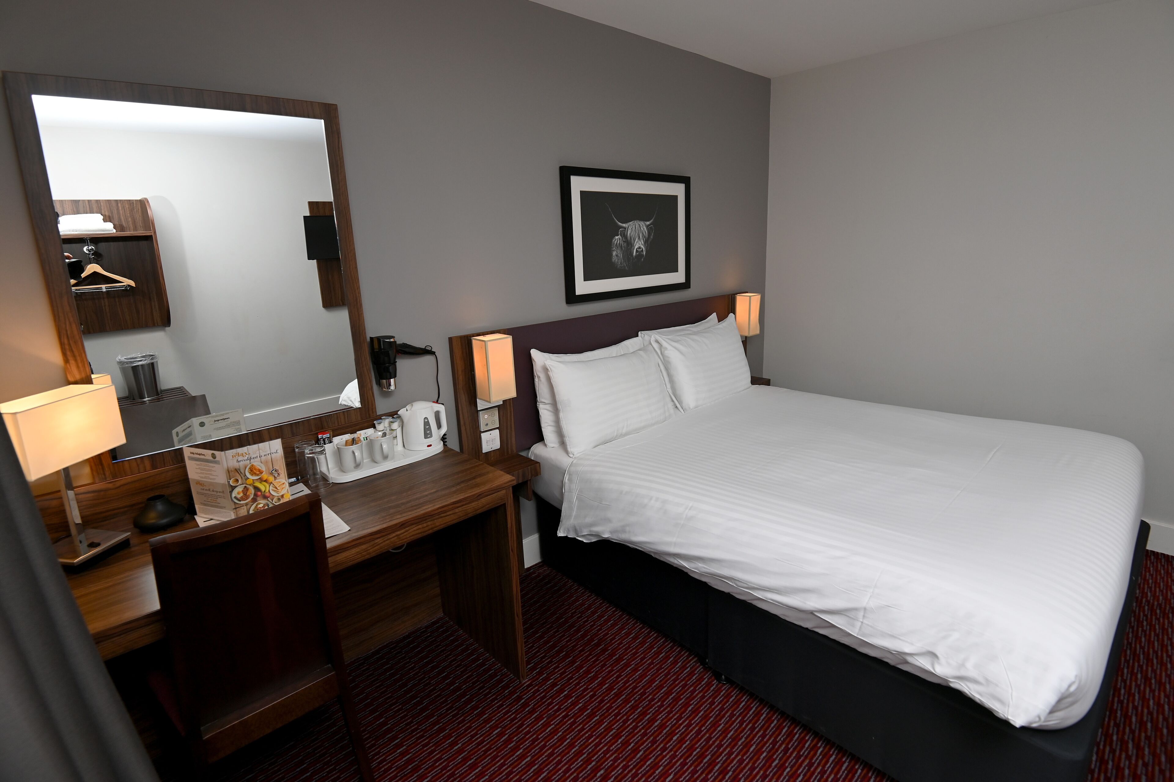 Habitación de hotel con cama, escritorio con lámpara, espejo, cuadro en la pared y alfombra roja.