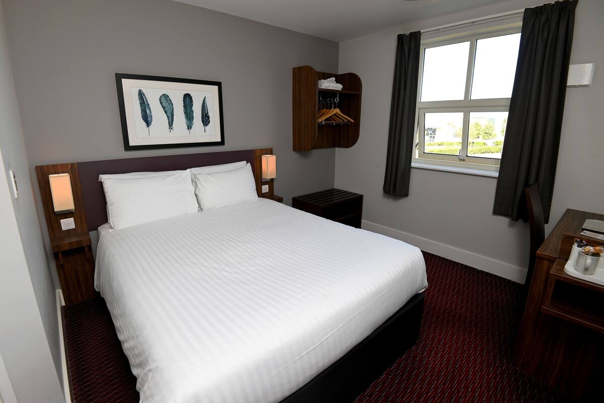 A hotel room with a large bed, a nightstand with a lamp, a wooden wardrobe, a window with curtains, and a framed artwork on the wall.