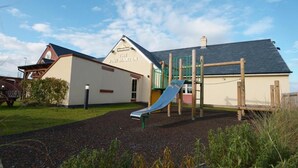 Un parque infantil con tobogán y estructuras para escalar, un edificio con el letrero 'The Pine Marten', y un cielo despejado.