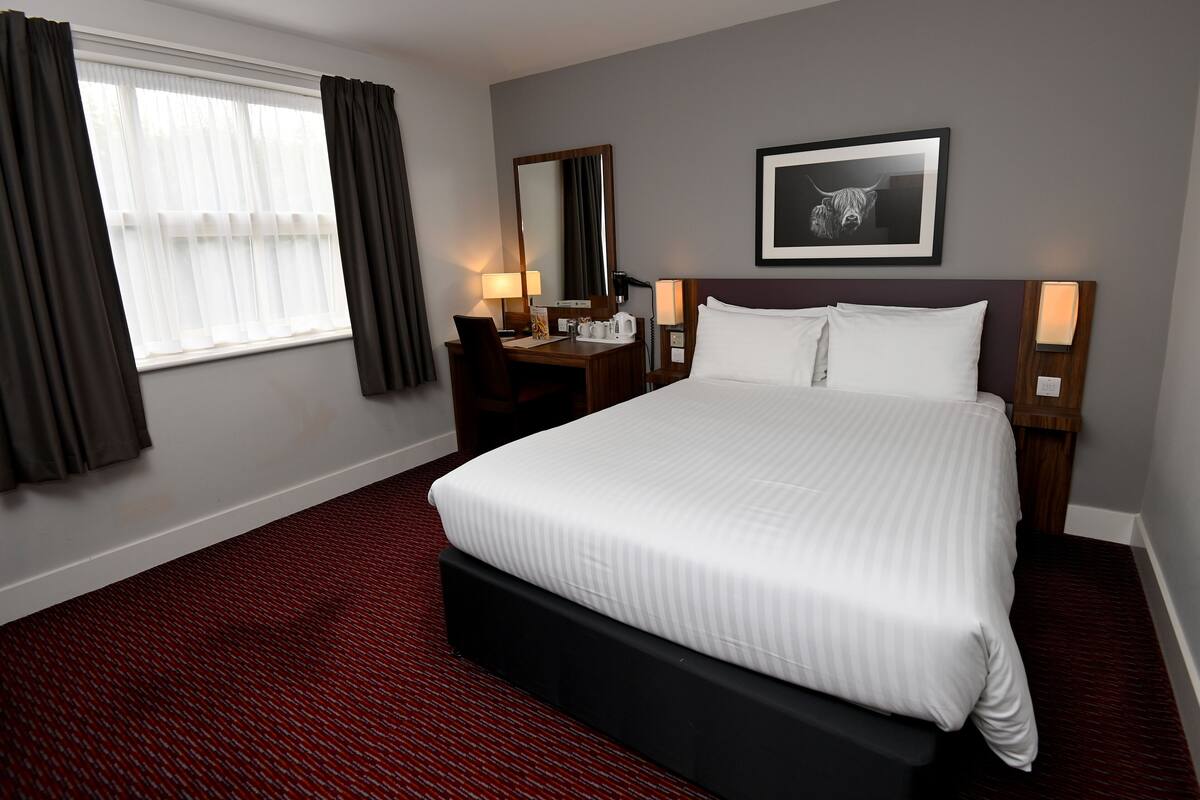 A hotel room with a large bed, a desk with a chair, a desk lamp, a framed picture on the wall, and a window with curtains.