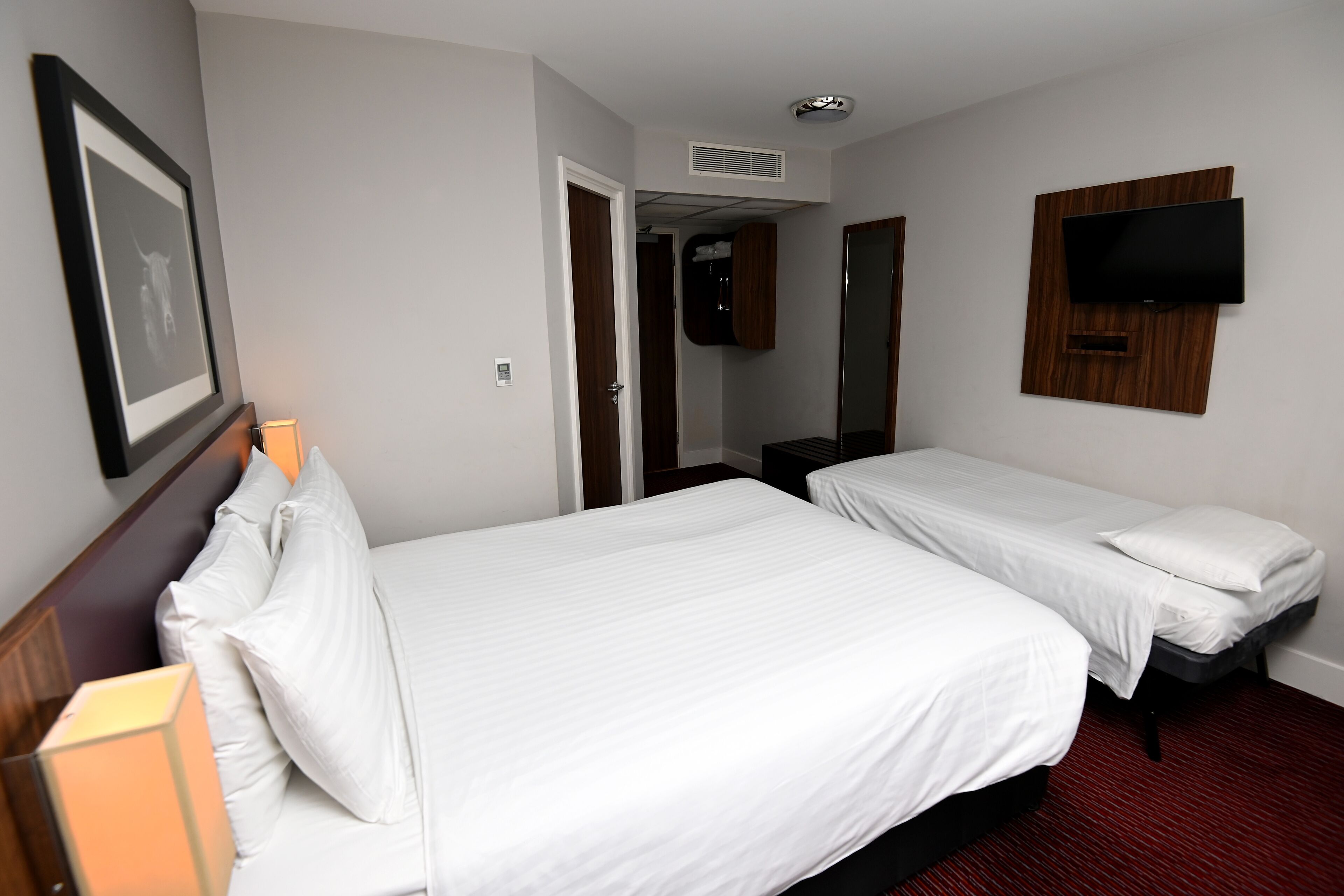 A hotel room with two single beds, a flat-screen TV mounted on the wall, a framed picture on the wall, and a door leading to another room.