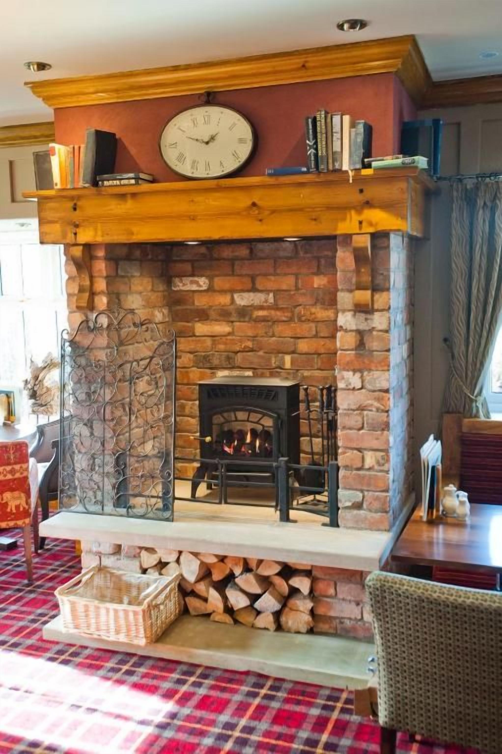 A fireplace with a clock above it, surrounded by books and a basket of firewood.