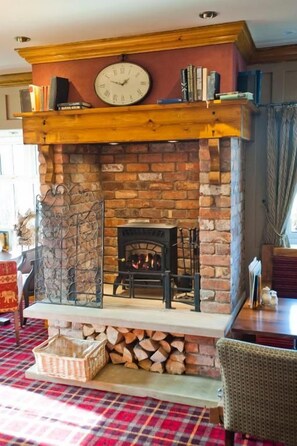 A fireplace with a clock above it, surrounded by books and a basket of firewood. - Pine Marten, Dunbar by Marston's Inns (Dunbar)