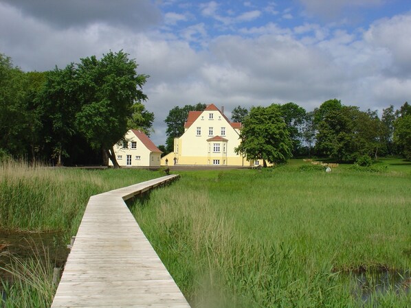 A wooden pathway leading to a yellow building with a red roof, surrounded by lush greenery and trees.