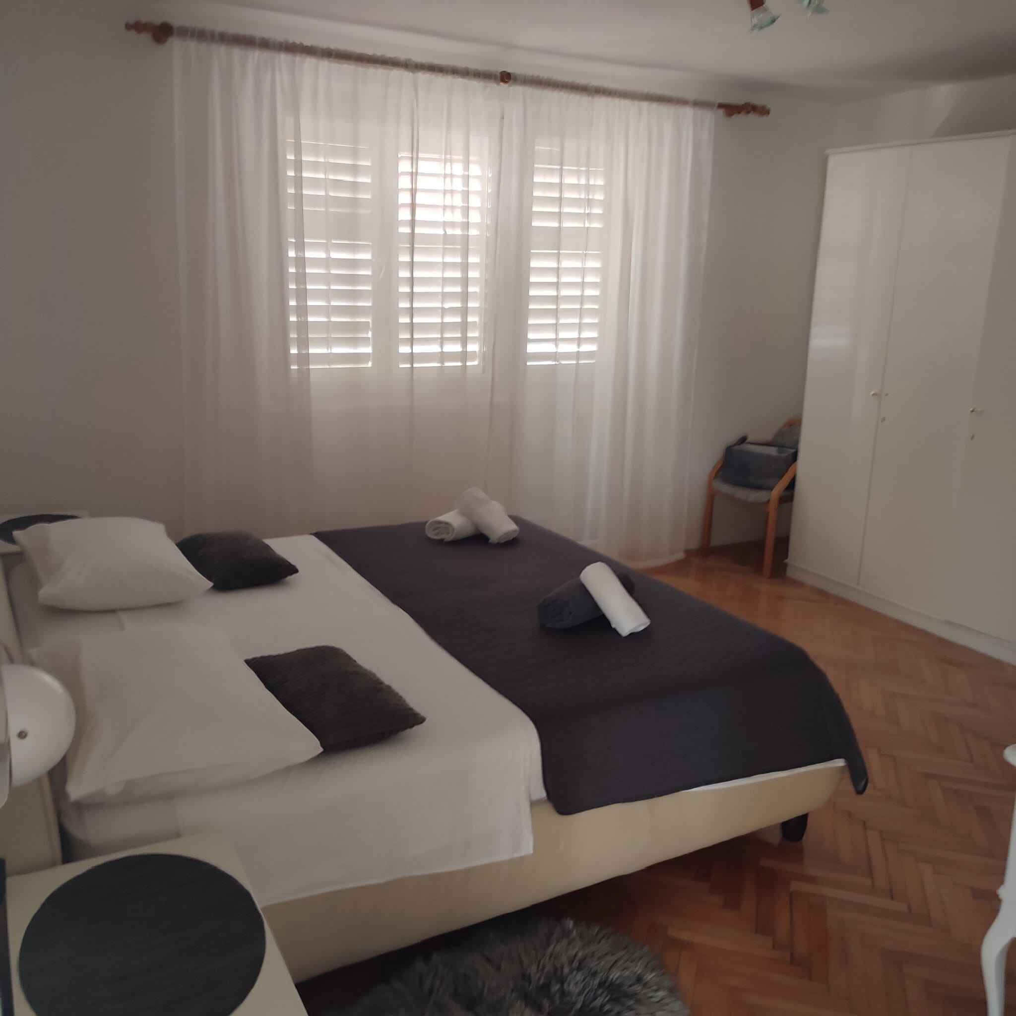 En pænt redt seng med puder og dyne, et natbord med lampe, et klædeskab og et vindue med skodder.