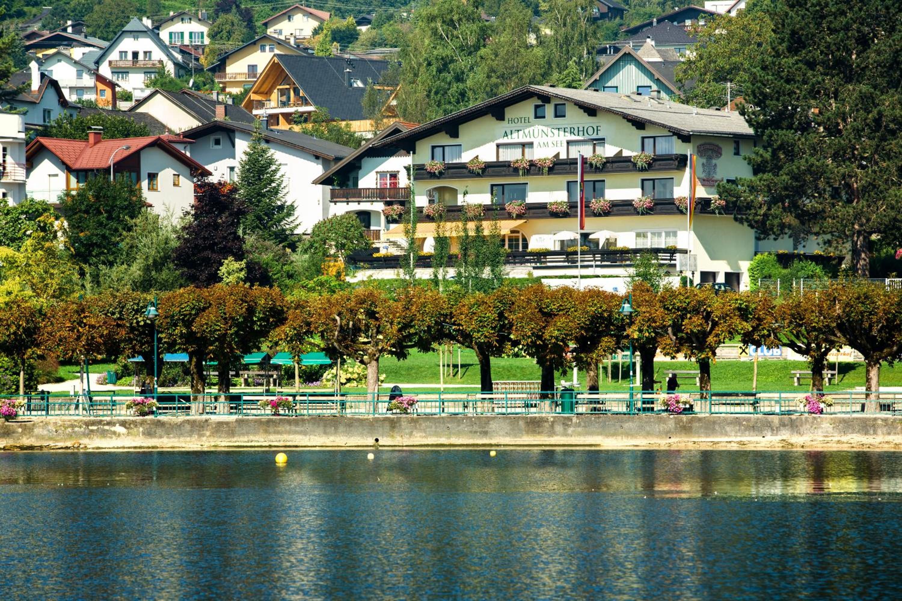 Ein Hotel im traditionellen alpinen Baustil, gelegen am Ufer eines ruhigen Sees mit einem Park im Vordergrund.