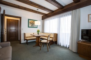 A room with a wooden ceiling, a dining table with chairs, a sofa, a television on a wooden cabinet, and a window with curtains.